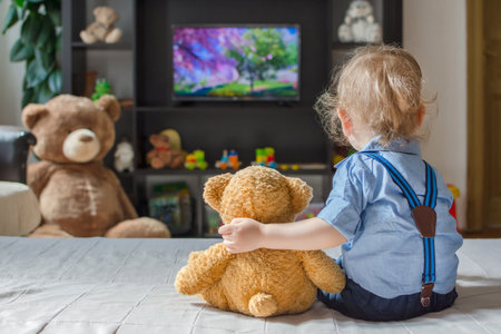Cute baby boy and his teddy bear watching TV sitting on a couch in the living room at homeの写真素材
