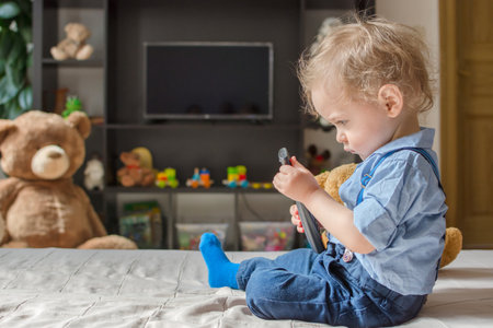 Cute baby boy playing with the remote control to watch TV sitting on a couch with his teddy bear, at homeの写真素材