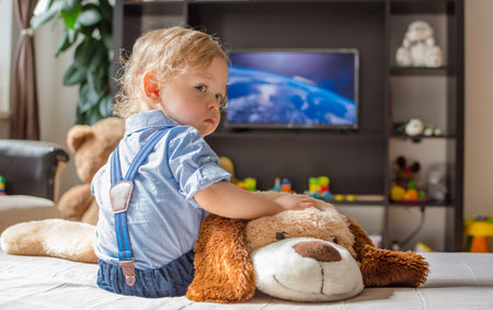 Cute baby boy and his dog plush toy watching TV sitting on a couch in the living room at homeの写真素材