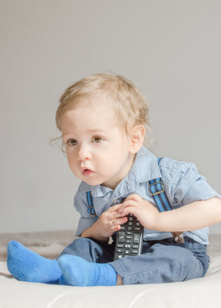 Cute baby boy playing with the remote control to watch TV sitting on a couch, at homeの写真素材