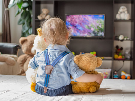 Cute baby boy and his teddy bear watching TV sitting on a couch in the living room at homeの写真素材