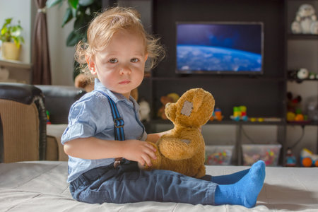 Cute baby boy and his teddy bear watching TV sitting on a couch in the living room at homeの写真素材