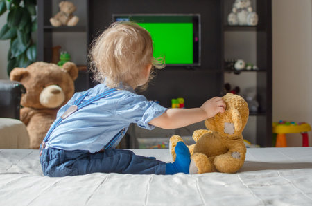 Cute baby boy and his teddy bear watching TV sitting on a couch in the living room at homeの写真素材