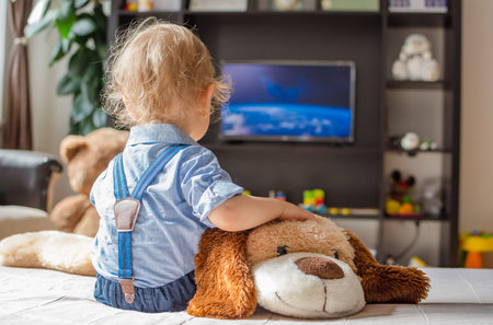 Cute baby boy and his dog plush toy watching TV sitting on a couch in the living room at homeの写真素材