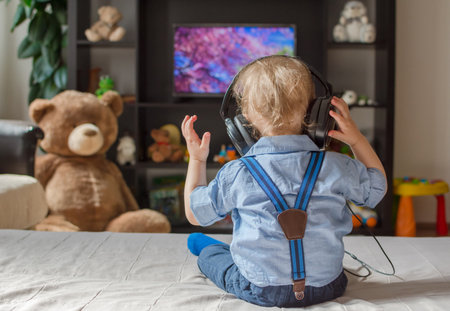 Cute baby boy with headphones watching TV sitting on a couch in the living room at homeの写真素材