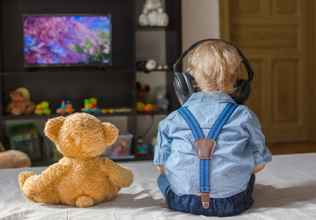 Cute baby boy with headphones and his teddy bear watching TV sitting on a couch in the living room at homeの写真素材