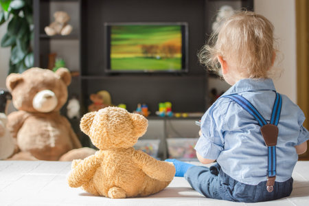 Cute baby boy and his teddy bear watching TV sitting on a couch in the living room at homeの写真素材