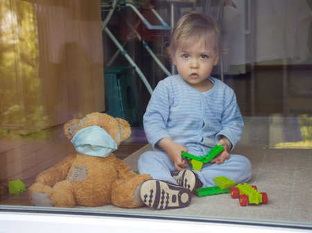 Child in home quarantine playing at the window with sick teddy bear wearing a medical mask against viruses during coronavirus COVID-2019 and flu outbreak. Photo made through the windowの写真素材