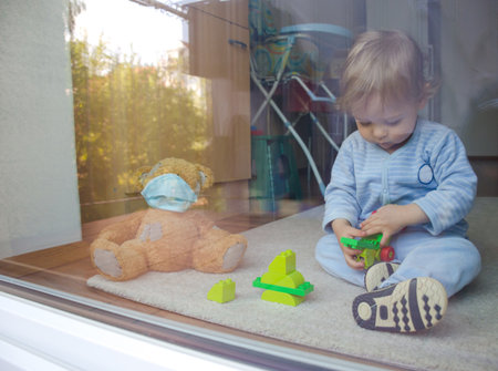 Child in home quarantine playing at the window with sick teddy bear wearing a medical mask against viruses during coronavirus COVID-2019 and flu outbreak. Photo made through the windowの写真素材