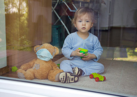 Child in home quarantine playing at the window with sick teddy bear wearing a medical mask against viruses during coronavirus COVID-2019 and flu outbreak. Photo made through the windowの写真素材