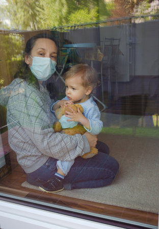 Mother and child in home quarantine playing at the window with sick teddy bear wearing a medical mask against viruses during coronavirus COVID-2019 and flu outbreak. Photo made through the windowの写真素材