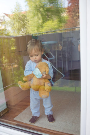 Child in home quarantine playing at the window with sick teddy bear wearing a medical mask against viruses during coronavirus COVID-2019 and flu outbreak. Photo made through the windowの写真素材