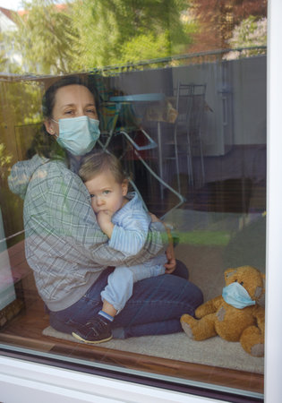 Mother and child in home quarantine staying at the window with sick teddy bear wearing a medical mask against viruses during coronavirus COVID-2019 and flu outbreak. Photo made through the windowの写真素材