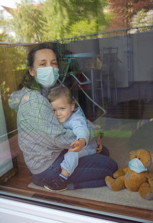 Mother and child in home quarantine staying at the window with sick teddy bear wearing a medical mask against viruses during coronavirus COVID-2019 and flu outbreak. Photo made through the windowの写真素材