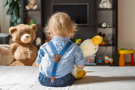 Cute baby boy holding his plush toy watching TV sitting on a couch in the living room at homeの写真素材