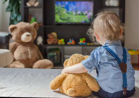 Cute baby boy and his teddy bear watching TV sitting on a couch in the living room at homeの写真素材