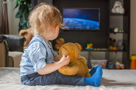 Cute baby boy and his teddy bear watching TV sitting on a couch in the living room at homeの写真素材