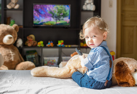 Cute baby boy and his dog plush toy watching TV sitting on a couch in the living room at homeの写真素材