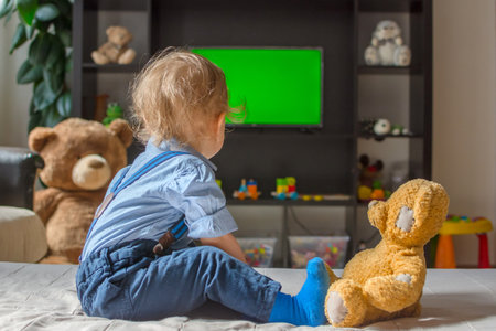 Cute baby boy and his teddy bear watching TV sitting on a couch in the living room at homeの写真素材