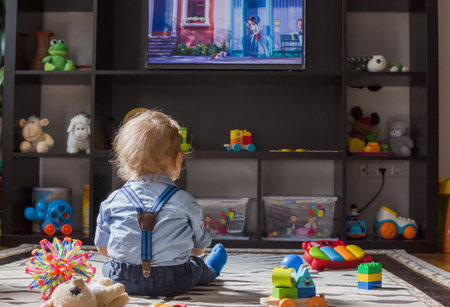 Cute baby boy and his teddy bear watching TV sitting on the floor covered with toys, at homeの写真素材