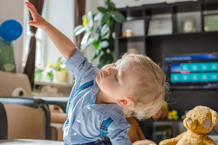Cute baby boy and his teddy bear watching TV sitting on the floor covered with toys, at homeの写真素材