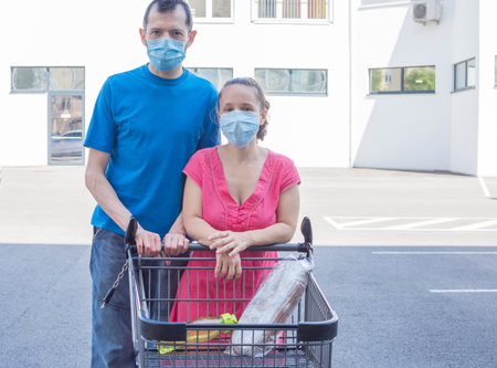 Couple wearing medical masks standing with an almost empty shopping cart in front of a store during coronavirus pandemic. Concept of economic crisis affecting people due to COVID-2019 diseaseの写真素材