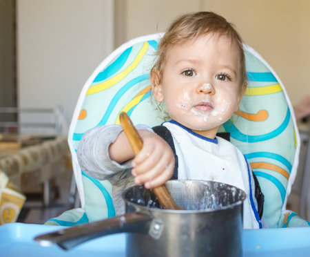 Funny baby child getting messy eating cereals or porridge by itself with a wooden spoon, straight from the cooking potの写真素材