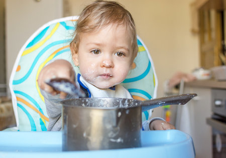 Funny baby child getting messy eating cereals or porridge by itself with a wooden spoon, straight from the cooking potの写真素材