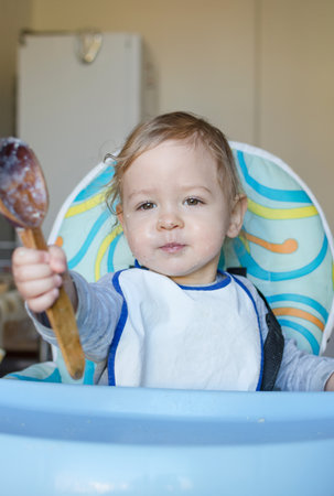 Funny baby child getting messy eating cereals or porridge by itself with a wooden spoon, straight from the cooking potの写真素材