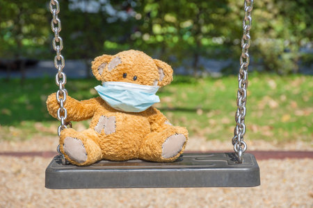 A lonely little teddy bear wearing a medical mask, sitting on a swing in the park on a beautiful sunny day. Empty children playground, illness COVID-2019 disease conceptの写真素材