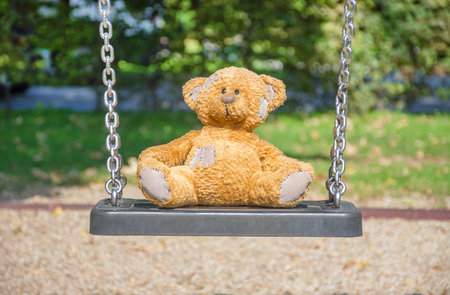 Cute little teddy bear standing alone on a swing in empty park on a beautiful sunny day.の写真素材
