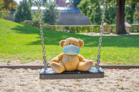 Lonely little teddy bear wearing a medical mask, sitting on a swing in the park on a beautiful sunny day. Empty children playground, illness COVID-2019 disease conceptの写真素材