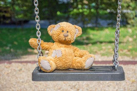 Cute little teddy bear standing alone on a swing in empty park on a beautiful sunny day.の写真素材