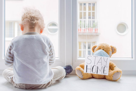 Child in home quarantine at the window putting a medical mask on his sick teddy bear, for protection against viruses during coronavirus COVID-19 and flu lock down.Children and illness disease conceptの写真素材