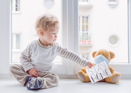 Child in home quarantine at the window putting a medical mask on his sick teddy bear, for protection against viruses during coronavirus COVID-19 and flu lock down.Children and illness disease conceptの写真素材