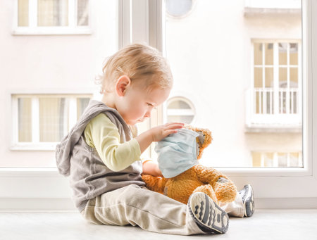 Child in home quarantine at the window putting a medical mask on his sick teddy bear, for protection against viruses during coronavirus COVID-19 and flu lock down.Children and illness disease conceptの写真素材