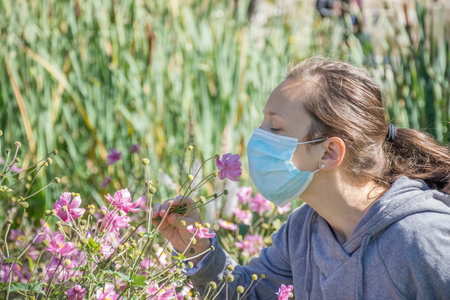 Young woman trying to smell wild flowers in the park while wearing medical mask. Conceptual photo of coronavirus or allergy, quarantine exit.の写真素材