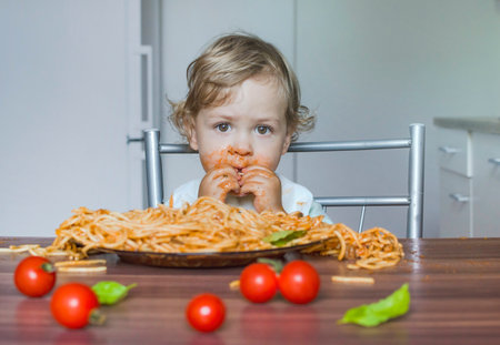 Funny baby child getting messy eating spaghetti with tomato sauce from a large plate, by itself with his hands, at homeの写真素材