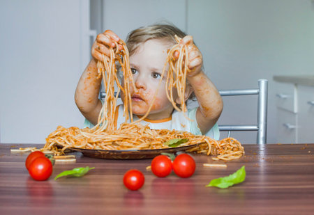 Funny baby child getting messy eating spaghetti with tomato sauce from a large plate, by itself with his hands, at homeの写真素材
