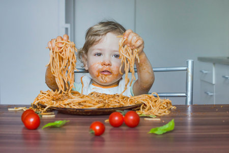 Funny baby child getting messy eating spaghetti with tomato sauce from a large plate, by itself with his hands, at homeの写真素材