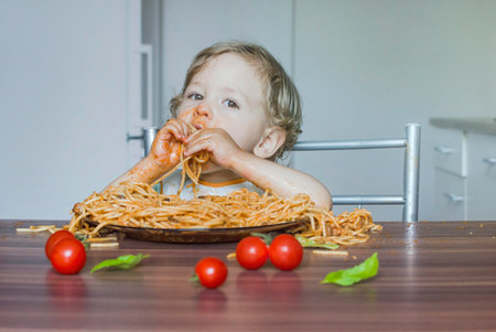 Funny baby child getting messy eating spaghetti with tomato sauce from a large plate, by itself with his hands, at homeの写真素材