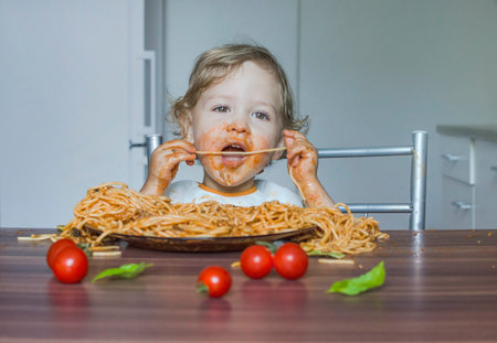 Funny baby child getting messy eating spaghetti with tomato sauce from a large plate, by itself with his hands, at homeの写真素材