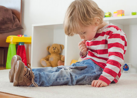 Cute little child using a smartphone while sitting on the floor at home. Digital device and screen time addiction, technology in the hands of children concept.の写真素材