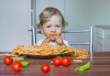 Funny baby child getting messy eating spaghetti with tomato sauce from a large plate, by itself with his hands, at homeの写真素材