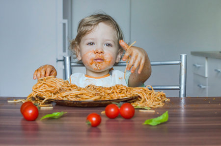 Funny baby child getting messy eating spaghetti with tomato sauce from a large plate, by itself with his hands, at homeの写真素材