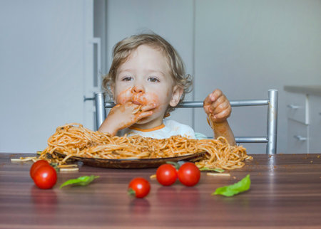 Funny baby child getting messy eating spaghetti with tomato sauce from a large plate, by itself with his hands, at homeの写真素材