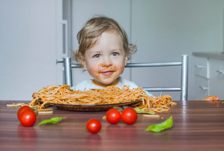 Funny baby child getting messy eating spaghetti with tomato sauce from a large plate, by itself with his hands, at homeの写真素材