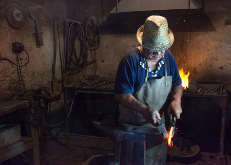 Salaj, Romania-May 12, 2018: Old blacksmith working hot metal horseshoe with hammer on the anvil in his vintage workshop in Transylvania region, Romaniaのeditorial素材