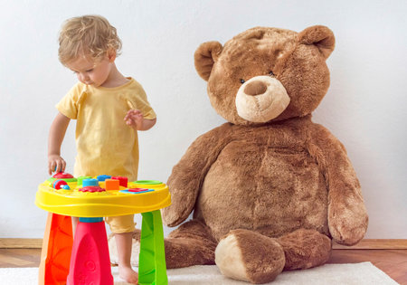 Sweet happy child boy having fun playing at a colorful table of activities with his giant teddy bear, indoor at homeの写真素材