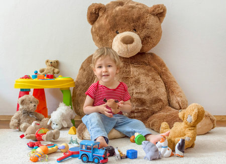 Sweet happy child boy having fun playing with his giant teddy bear and many colorful toys, indoor at homeの写真素材